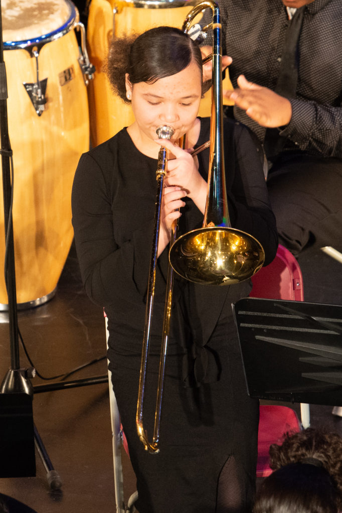 A young woman with light skin and a puffy pony tail plays trombone during a performance at the Strand Theatre in 2019