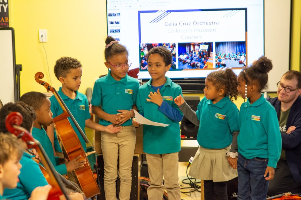 Image of students wearing teal shirts with logo at a school event. Some students are presenting work, and others have cellos.