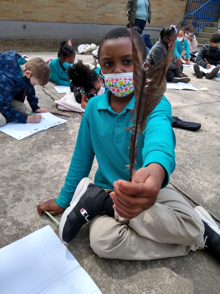 A student in first grade who is black holds up a feather that he is drawing during fieldwork