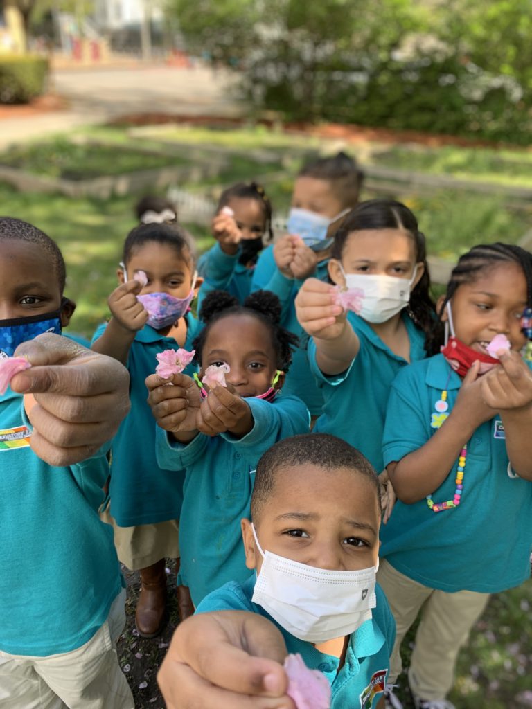 A group of students in Pre-K with teal shirts hold up flowers. They are wearing masks because the photo was taken in the spring of 2021.